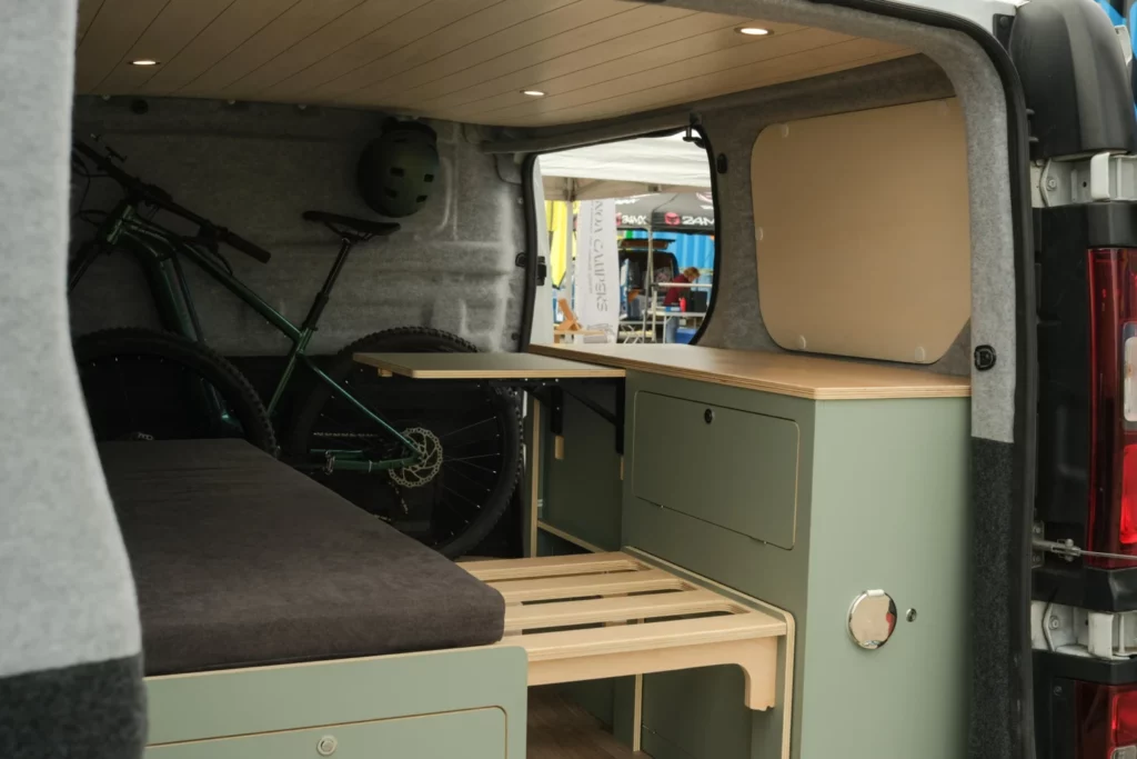 Van aménagé par Avanoa Campers avec rangement vélos, module cuisine vert sauge et lit, sur le stand du Provence Van Week-End