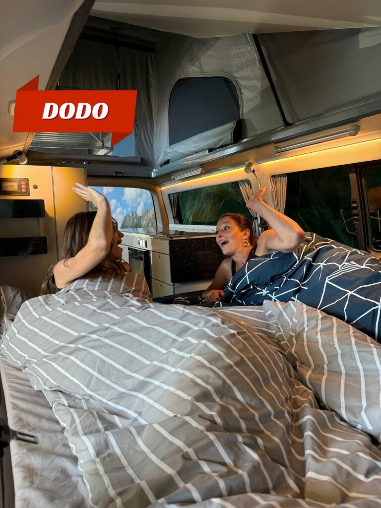 Deux amies allongées dans le couchage double d'un van aménagé REIMO Eco Campers, ambiance conviviale et rieuse