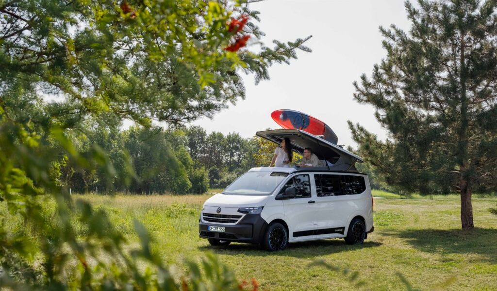 Van aménagé Volkswagen Triostyle REIMO garé en forêt, toit relevable ouvert avec paddle sur le toit, deux personnes à l'intérieur