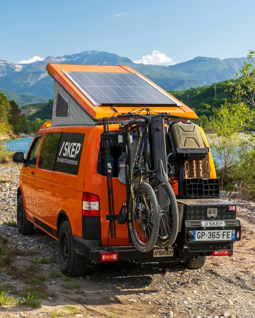 Van orange SKEP équipé d'un porte-vélos arrière et panneau solaire sur toit relevable, montagnes en arrière-plan