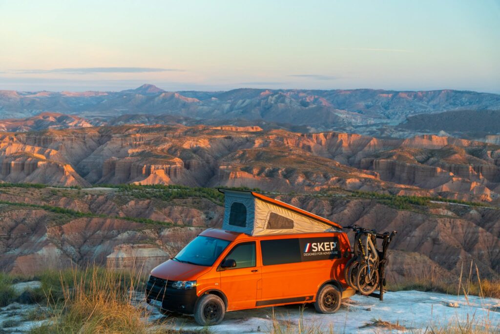 Van orange SKEP avec toit relevable ouvert et porte-vélos, garé face à un paysage de canyons au coucher du soleil
